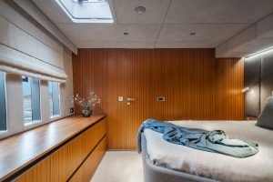 Interior view of a Sunreef Yachts bedroom featuring a neatly made bed with a blue throw on it, wooden paneled walls, a row of windows on the left with closed blinds, and a dresser with a vase of decorative branches.