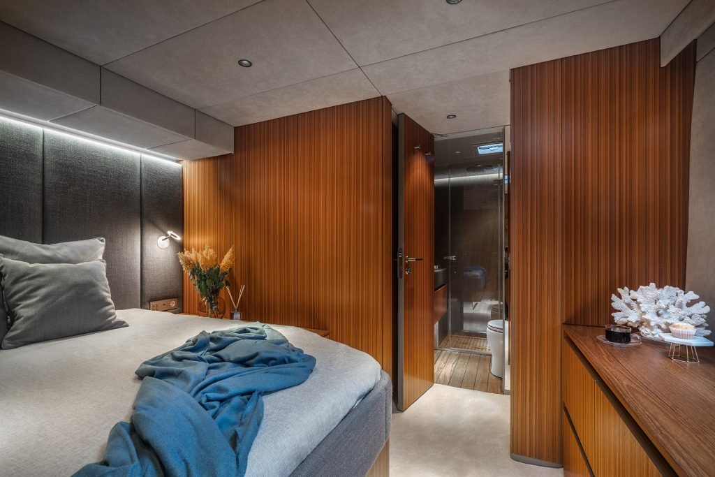A luxurious bedroom interior on a Sunreef Yachts vessel. The room features a cozy bed with gray and light blue bedding, a wooden wall with a door leading to an en-suite bathroom. A decorative coral and vase with dried flowers are on a wooden dresser.