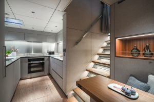 A luxurious interior of a Sunreef Yachts vessel featuring a modern kitchenette, sleek cabinetry, and wooden steps leading to the upper level. The space is well-lit and elegantly designed, with a small dining area.