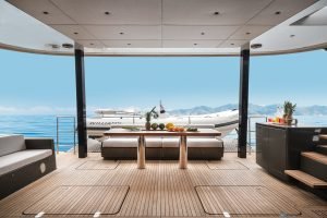 A luxury yacht deck by Sunreef Yachts features a modern interior with wooden flooring. In the background, a sleek inflatable boat is seen against a backdrop of clear blue water and mountains.