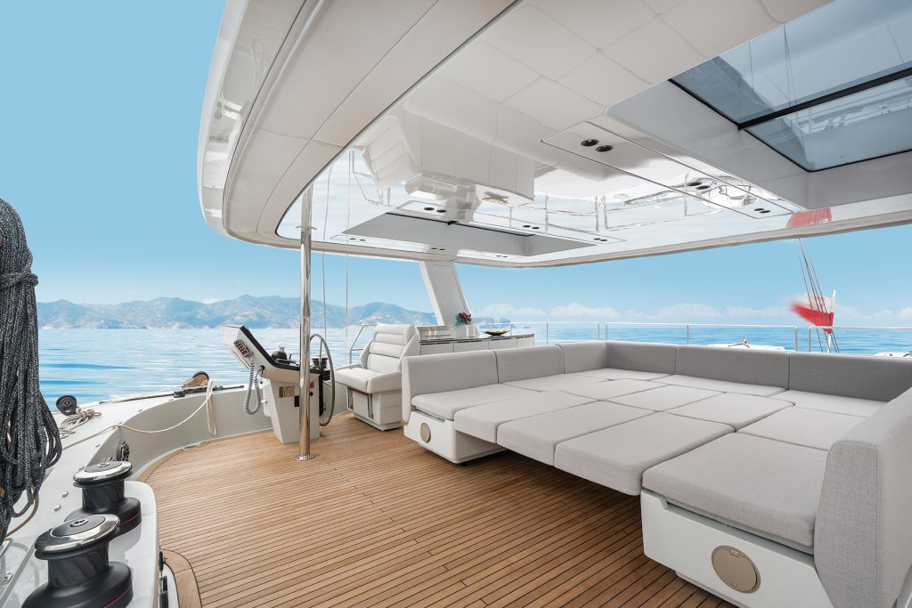 The image shows the deck of a Sunreef Yachts luxury catamaran, featuring spacious seating and a control panel with a view of the ocean and distant mountains under a clear blue sky.