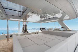 An interior view of a Sunreef Yachts catamaran, featuring a spacious lounge area with beige cushioned seating under a sleek, modern roof. The ocean is visible through large windows, with mountains in the background.