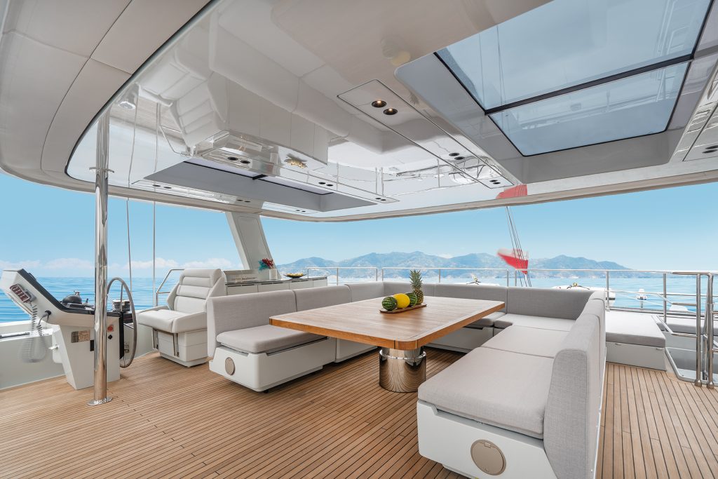 Luxurious interior of a Sunreef Yachts vessel featuring a spacious seating area with a wooden table and cushioned benches. A panoramic view of the ocean and distant mountains is visible through the large windows and skylights.