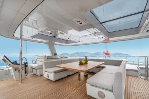 Luxurious interior of a Sunreef Yachts vessel featuring a spacious seating area with a wooden table and cushioned benches. A panoramic view of the ocean and distant mountains is visible through the large windows and skylights.