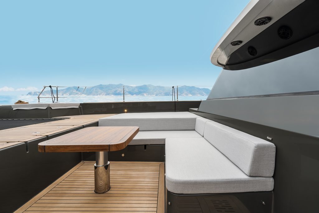 The image features the deck of a luxury yacht by Sunreef Yachts, showcasing a sleek and modern design with a wooden table and bench seating. The open sea and a clear blue sky are visible in the background.