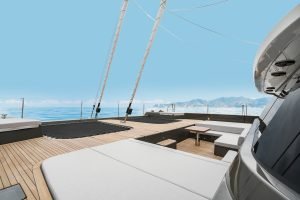 The deck of a luxury catamaran by Sunreef Yachts features spacious lounging areas with cushioned seating and a trampoline. The yacht is sailing on calm waters with mountains in the background under a clear blue sky.