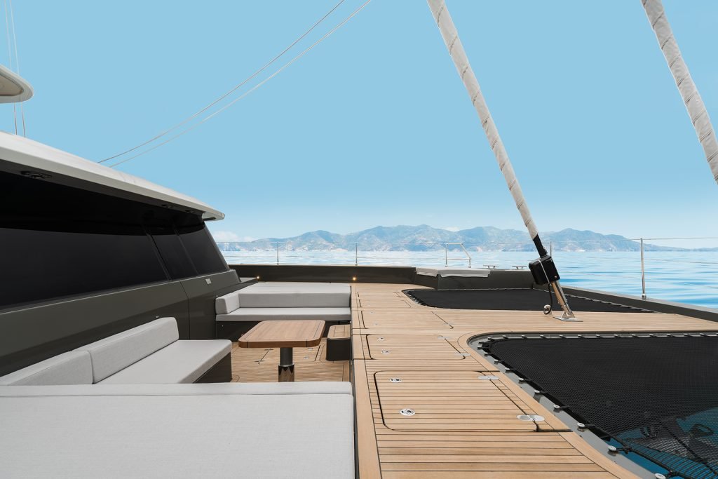 The image shows a luxurious deck of a Sunreef Yachts catamaran with modern seating, a wooden floor, and a table. It overlooks a calm sea with mountains in the background under a clear blue sky.