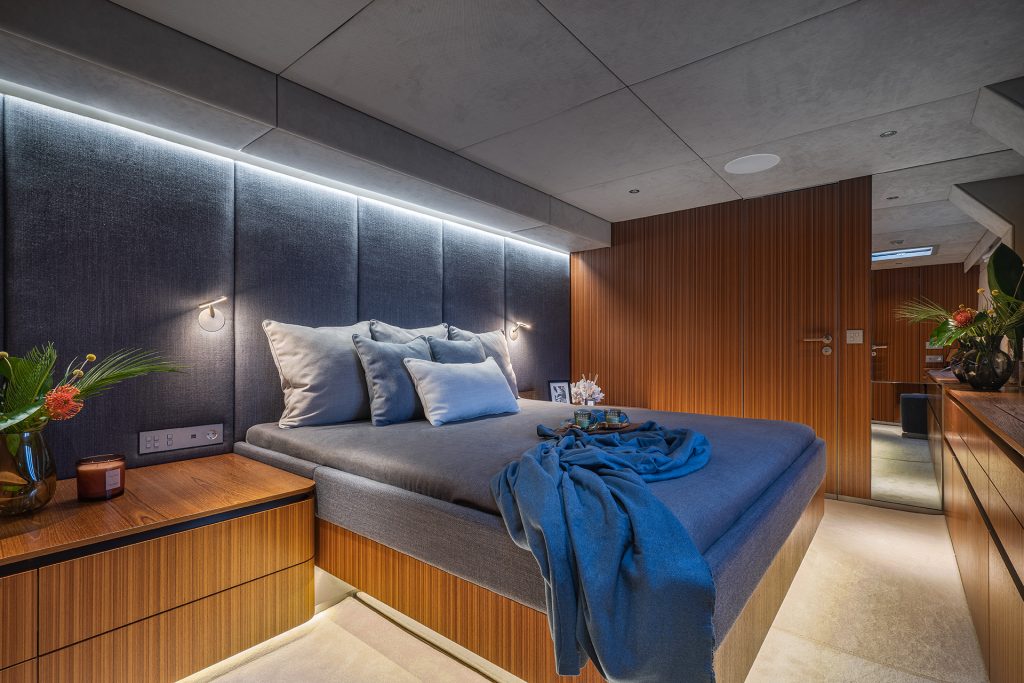 A luxurious bedroom interior on a Sunreef Yachts vessel featuring a large bed with grey and blue bedding, complemented by wood paneling and gentle lighting.