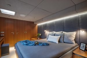 Interior of a luxury bedroom on a Sunreef Yachts vessel, featuring a large bed with gray bedding and blue throw, wooden paneling, and modern lighting.