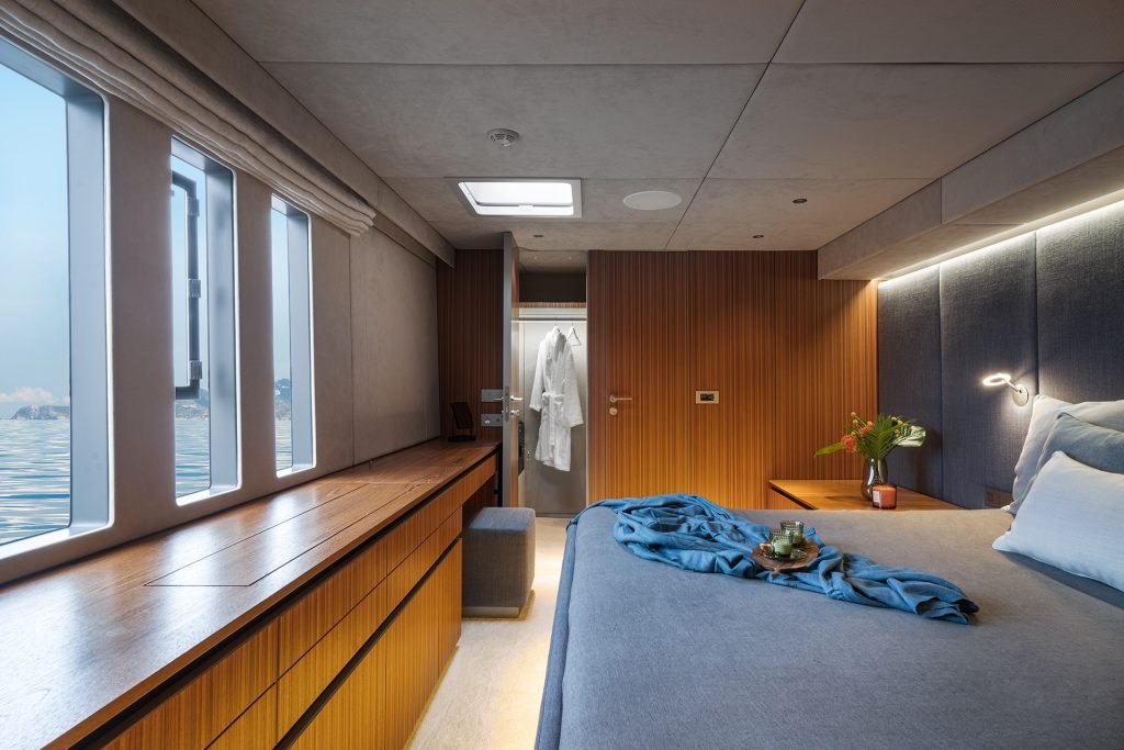The luxurious interior of a Sunreef Yachts cabin features a large bed with a blue throw, a wooden dresser, windows overlooking the sea, and a closet with a robe inside.