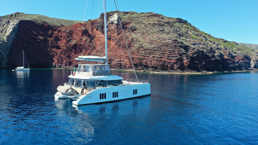 A Sunreef Yachts catamaran is anchored on calm blue waters near a rugged coastline with red and green cliffs. Another sailboat is visible in the distance.