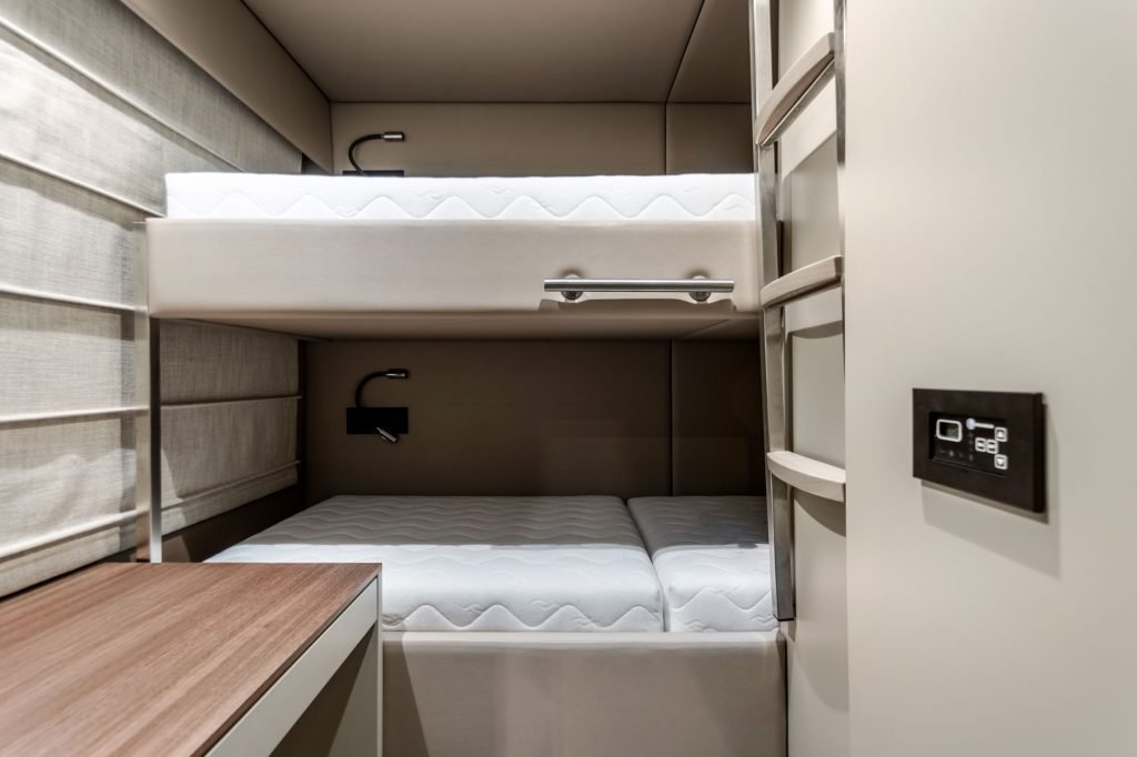 The image shows an interior of a Sunreef Yachts cabin featuring bunk beds with a sleek design. The beds have white mattresses and a modern aesthetic. The cabin walls are in neutral tones, with a wooden accent on one side. A control panel is visible on the right.