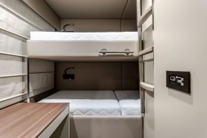 The image shows an interior of a Sunreef Yachts cabin featuring bunk beds with a sleek design. The beds have white mattresses and a modern aesthetic. The cabin walls are in neutral tones, with a wooden accent on one side. A control panel is visible on the right.