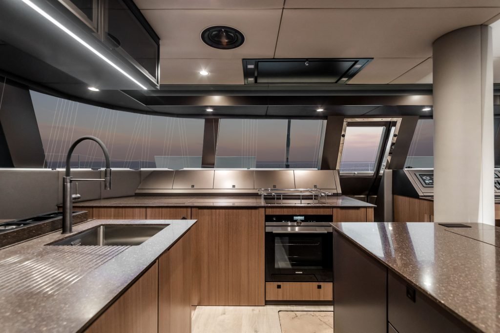 The image shows the luxurious interior of a Sunreef Yachts kitchen with modern appliances, wood cabinets, and a smooth countertop. Large windows offer a view of the sunset.