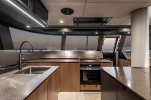 The image shows the luxurious interior of a Sunreef Yachts kitchen with modern appliances, wood cabinets, and a smooth countertop. Large windows offer a view of the sunset.