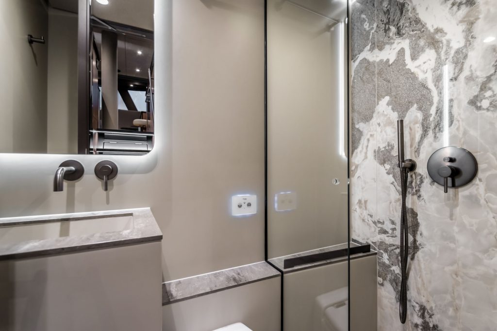 The image shows a modern bathroom interior on a Sunreef Yachts vessel. It features sleek grey and black surfaces, a large mirror, a minimalist sink, and a stylish shower area with marble-like wall patterns.