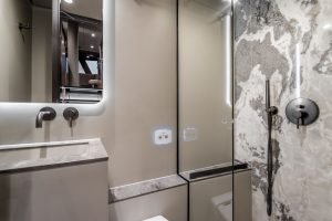 The image shows a modern bathroom interior on a Sunreef Yachts vessel. It features sleek grey and black surfaces, a large mirror, a minimalist sink, and a stylish shower area with marble-like wall patterns.