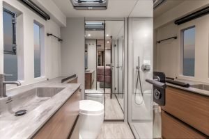 The image shows a luxurious bathroom on a Sunreef Yachts vessel, featuring modern design with marble countertops and sleek fixtures. Large mirrors and a glass shower complement the space, offering a view of the sea through the windows.