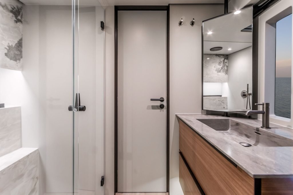 The interior of a modern bathroom on a Sunreef Yachts vessel, featuring a sleek design with a wooden vanity, a large mirror, and a glass-enclosed shower, with a view of the ocean through a window.