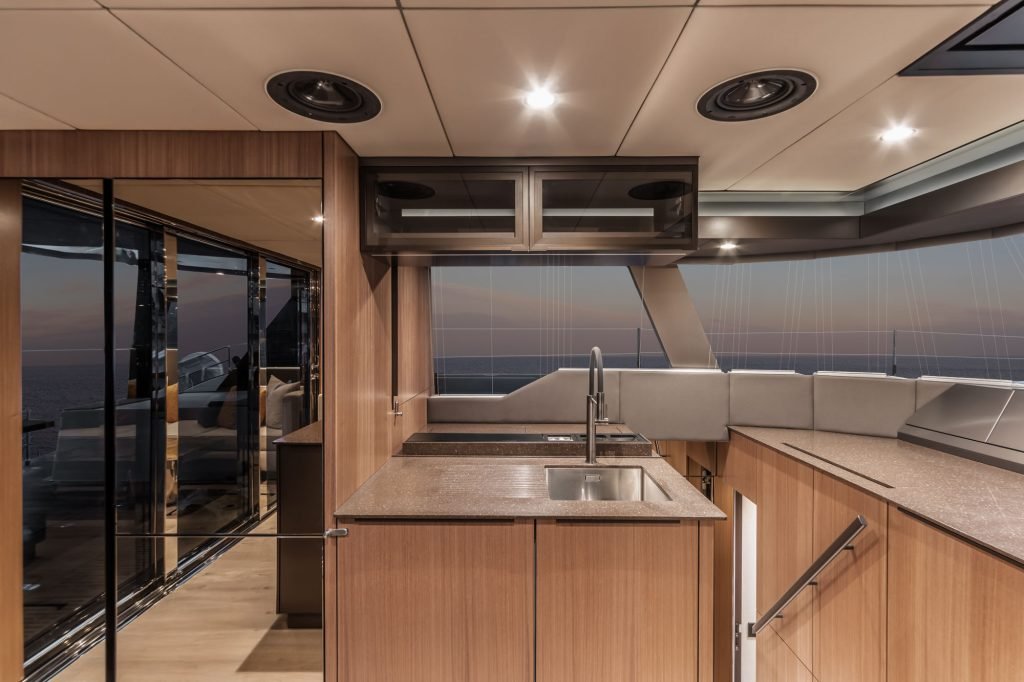 The interior of a Sunreef Yachts boat features a modern kitchen with wooden cabinets, a sink, and a countertop. Large windows provide a view of the ocean and a sunset sky.