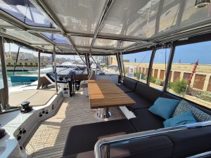 The interior of a Sunreef Yachts vessel featuring a spacious deck with a wooden table, cushioned seating, and protective canopy. The setting is bright with pillows for comfort.