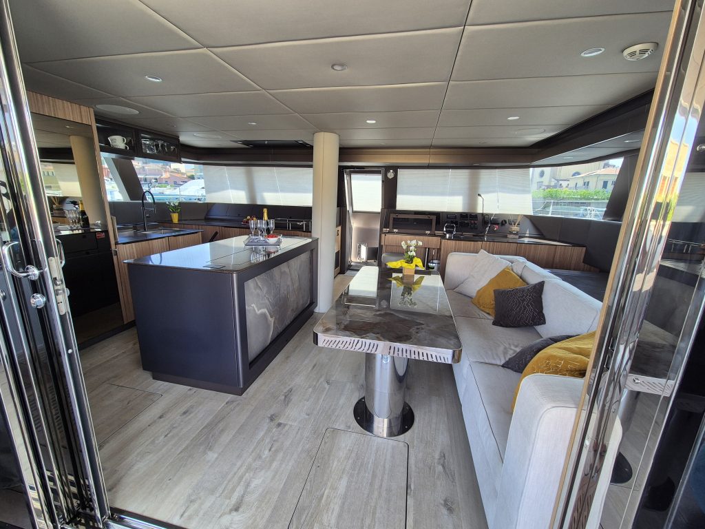 Interior of a Sunreef Yachts vessel featuring a modern living area with a sleek kitchen, a comfortable seating area with cushions on a sofa, a glass coffee table, and large windows.