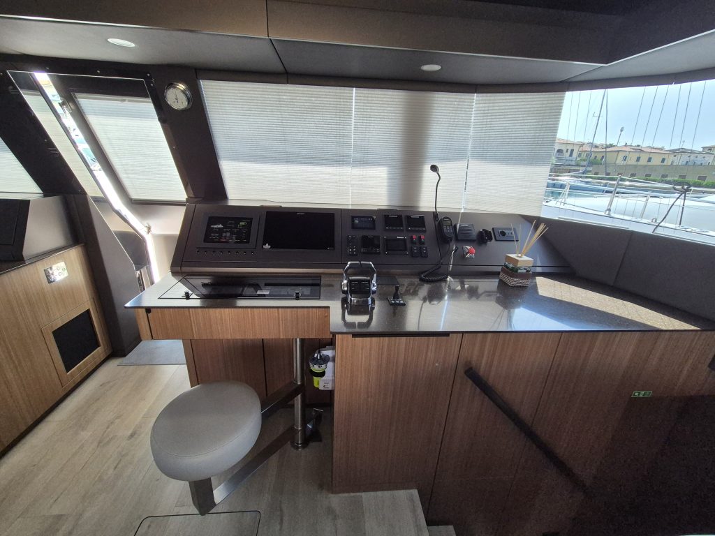 The cockpit of a Sunreef Yachts vessel featuring a polished wooden dashboard with various control panels and a stool. Large windows offer a view of the surroundings.