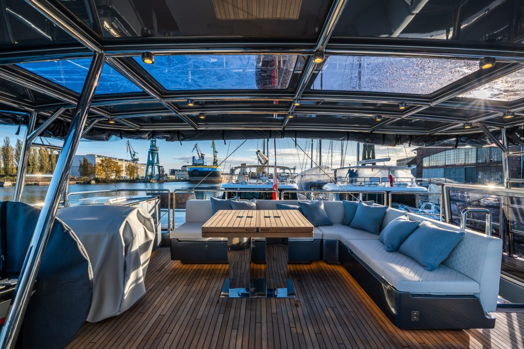 The Sunreef Yachts deck features a spacious seating area with a wooden table, surrounded by plush cushions. The backdrop includes a marina with various yachts, cranes, and industrial buildings under a blue sky.