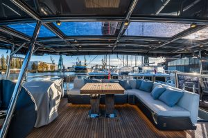 The Sunreef Yachts deck features a spacious seating area with a wooden table, surrounded by plush cushions. The backdrop includes a marina with various yachts, cranes, and industrial buildings under a blue sky.