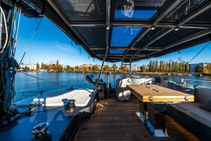 A luxurious deck of a Sunreef Yachts catamaran with a panoramic view of the water and distant cityscape, featuring seating areas and a wooden table under a glass roof.