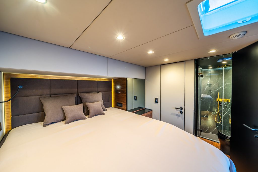 The image shows the interior of a Sunreef Yachts luxury cabin. The bedroom features a large bed with cushions, under soft ceiling lights. There is a modern bathroom visible through an open door, featuring a glass shower.