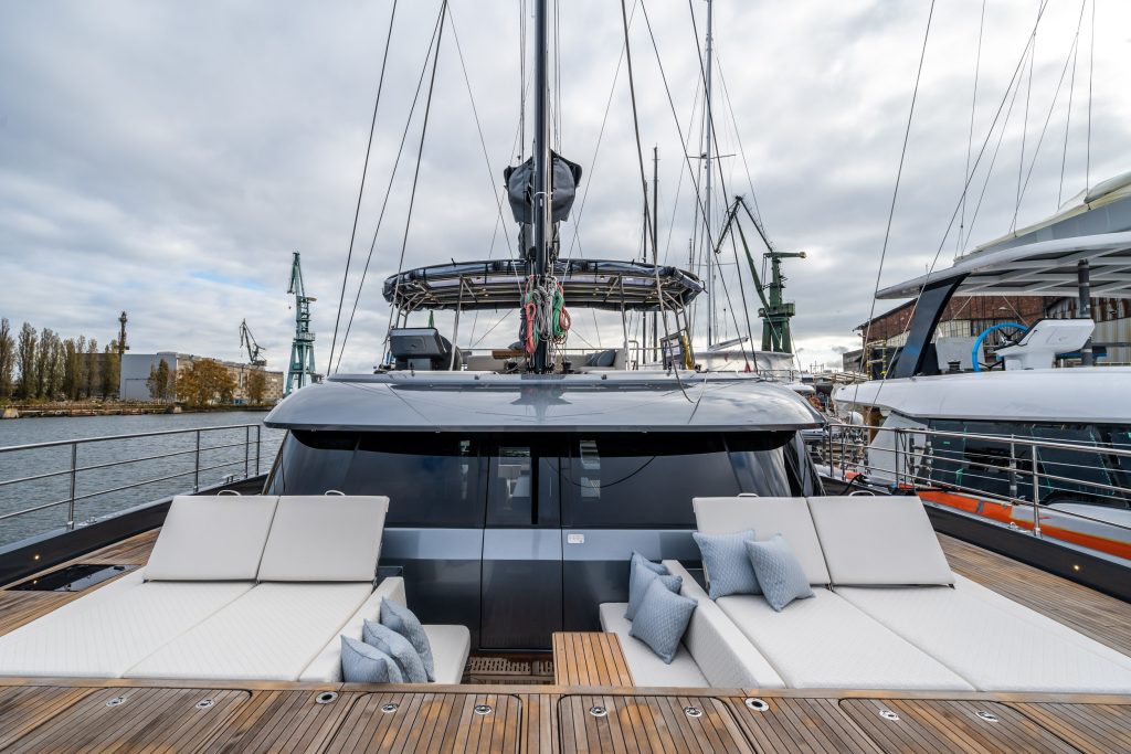 The image shows a luxury yacht by Sunreef Yachts docked at a marina. The deck features spacious seating areas with cushions, offering a view of nearby cranes and other boats against a cloudy sky.