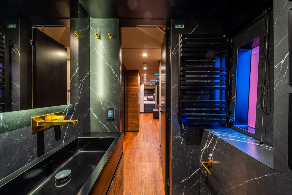Interior view of a luxurious bathroom on a Sunreef Yachts vessel, featuring dark marble walls, a black sink, and golden fixtures. A hallway with wooden flooring and modern decor is visible in the background.