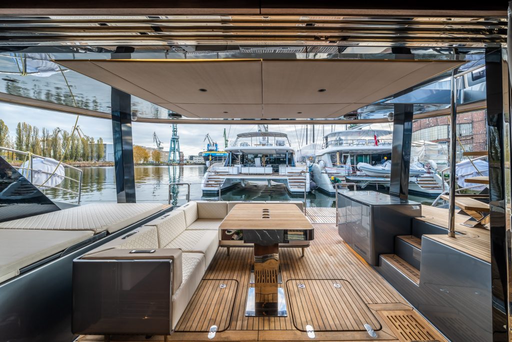 A view from the deck of a Sunreef Yachts luxury catamaran, showcasing a modern seating area with wooden flooring. Several other Sunreef Yachts are visible moored in the marina, with cranes and industrial buildings in the background.