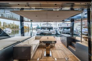 A view from the deck of a Sunreef Yachts luxury catamaran, showcasing a modern seating area with wooden flooring. Several other Sunreef Yachts are visible moored in the marina, with cranes and industrial buildings in the background.