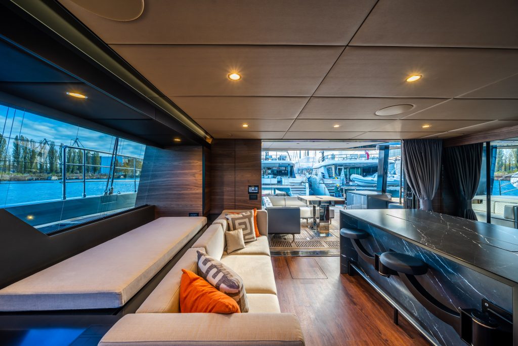 The interior of a Sunreef Yachts catamaran showcases a modern and luxurious design. There is a spacious lounge area with a large, comfortable sofa and colorful cushions. The room features a sleek bar counter with barstools, and large windows offer a view of the marina and nearby yachts.