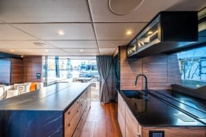 Interior view of a Sunreef Yachts luxury catamaran featuring a modern kitchen with a black countertop and wooden cabinets, large windows offering marina views, and a stylish seating area.
