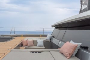 The deck of a Sunreef Yachts vessel features spacious seating with gray cushions and several decorative pillows. The surrounding sea and horizon are visible under a cloudy sky.