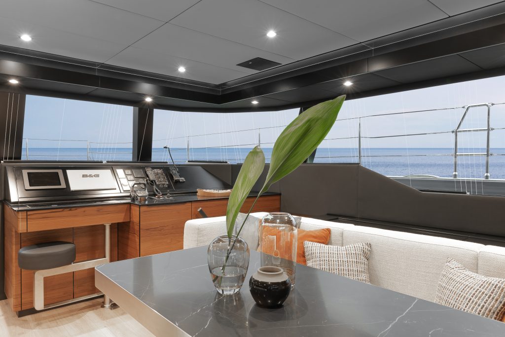 Interior of a luxury yacht featuring a navigation station with wooden cabinetry, a marble-topped table with decorative plants, and a view of the sea through large windows. The space is stylishly furnished with cushions, highlighting the high-end design typical of Sunreef Yachts.