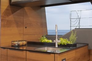 The image shows a modern kitchen interior on a Sunreef Yachts vessel. It features wooden cabinetry, a sleek black countertop with a built-in sink, and a view of the sea and sail rigging through a window. Fresh herbs in planters are placed on the countertop.