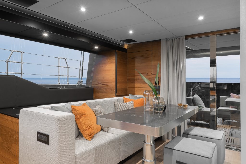 The interior of a luxurious Sunreef Yachts vessel features a modern dining area with a sleek metallic table, cushioned seating with orange and gray pillows, and decorative vases. The yacht has large windows offering a view of the sea.