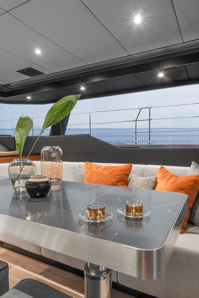 Interior view of a Sunreef Yachts vessel showing a modern and elegant dining area. The table is set with glasses and decorative items, surrounded by plush seating with orange cushions. Large windows offer a view of the sea.