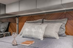 Interior of a Sunreef Yachts bedroom featuring a luxurious bed with a plush gray comforter, patterned pillows, a wooden headboard, and an hourglass along with a notebook beside it.