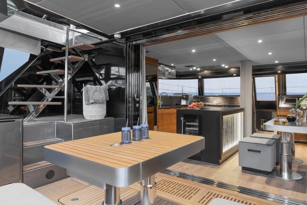 Interior view of a luxury yacht by Sunreef Yachts, featuring a modern dining area with a wooden table and blue glasses, adjacent to a sleek kitchen with a black island and modern appliances. There's a staircase leading to an upper deck.