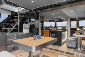 Interior view of a luxury yacht by Sunreef Yachts, featuring a modern dining area with a wooden table and blue glasses, adjacent to a sleek kitchen with a black island and modern appliances. There's a staircase leading to an upper deck.