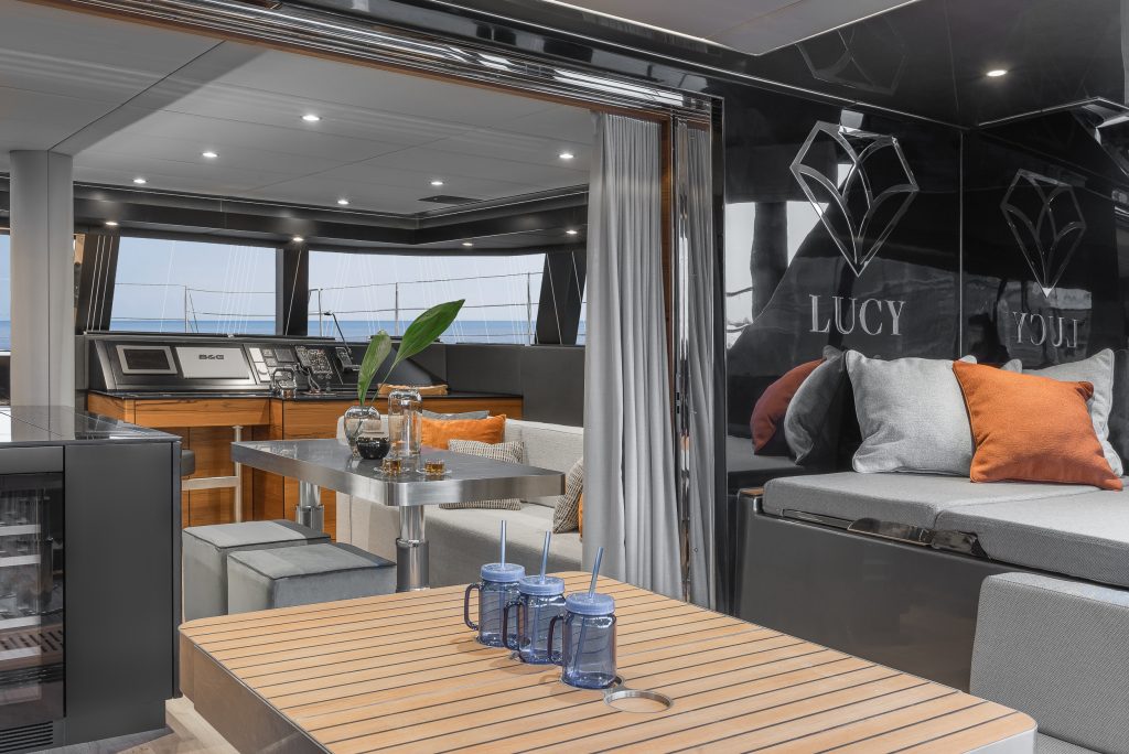Interior of a luxurious Sunreef Yachts catamaran with modern furnishings, including a dining area with a wooden table and cushioned seating, and a view of the helm and exterior deck.