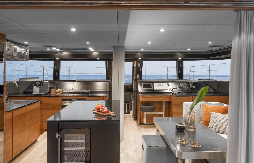 Interior of a luxurious yacht by Sunreef Yachts, featuring a modern kitchen with wooden cabinetry, a black countertop, a navigation area, and a sunlit lounging space with ocean views.
