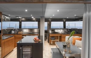 Interior of a luxurious yacht by Sunreef Yachts, featuring a modern kitchen with wooden cabinetry, a black countertop, a navigation area, and a sunlit lounging space with ocean views.