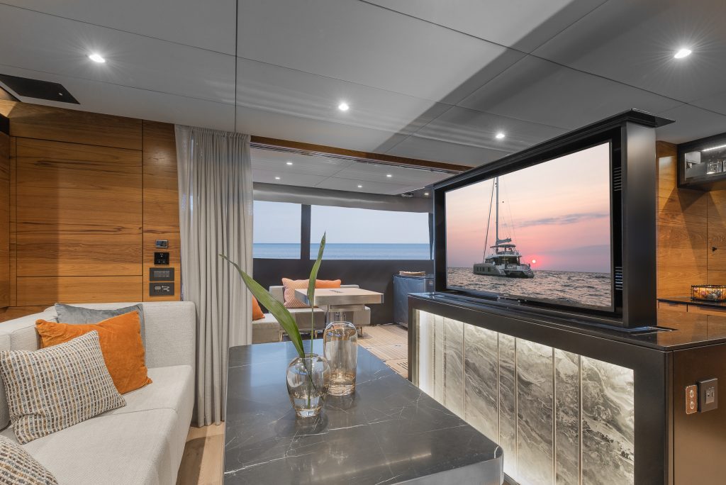 Interior of a luxurious Sunreef Yachts vessel featuring a plush seating area, wooden paneling, a large screen displaying a yacht at sunset, and modern decor elements.