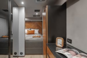 The interior of a Sunreef Yachts cabin features a modern design with a comfortable bed adorned with orange pillows and a throw blanket. The walls and furnishings are a blend of wood and sleek finishes, with a bedside table holding a framed photo and an open magazine.